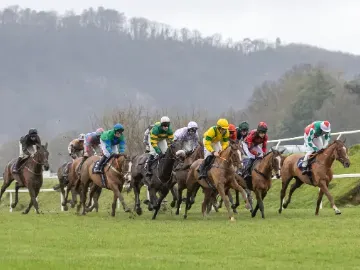 Horses and jockeys in colorful silks racing on the turf at Chepstow Racecourse with hills in the background.