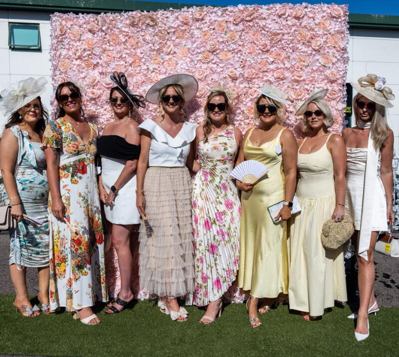 A large group of ladies dressed for the races pose in front of the flower wall at Chepstow