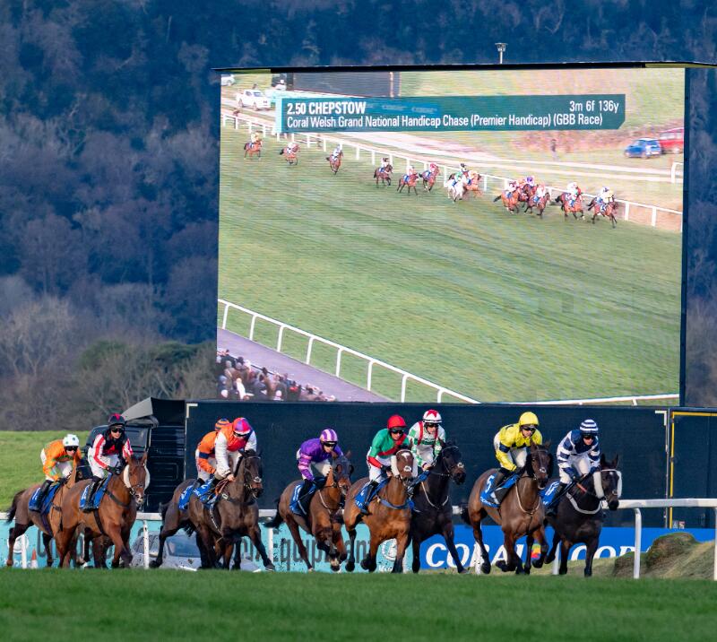 Horses race past a large viewing screen at Chepstow Races