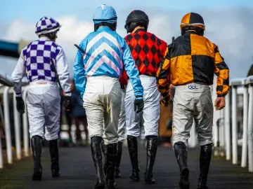 Four jockeys in colorful silks walking down the Chepstow Racecourse trackside walkway before a race